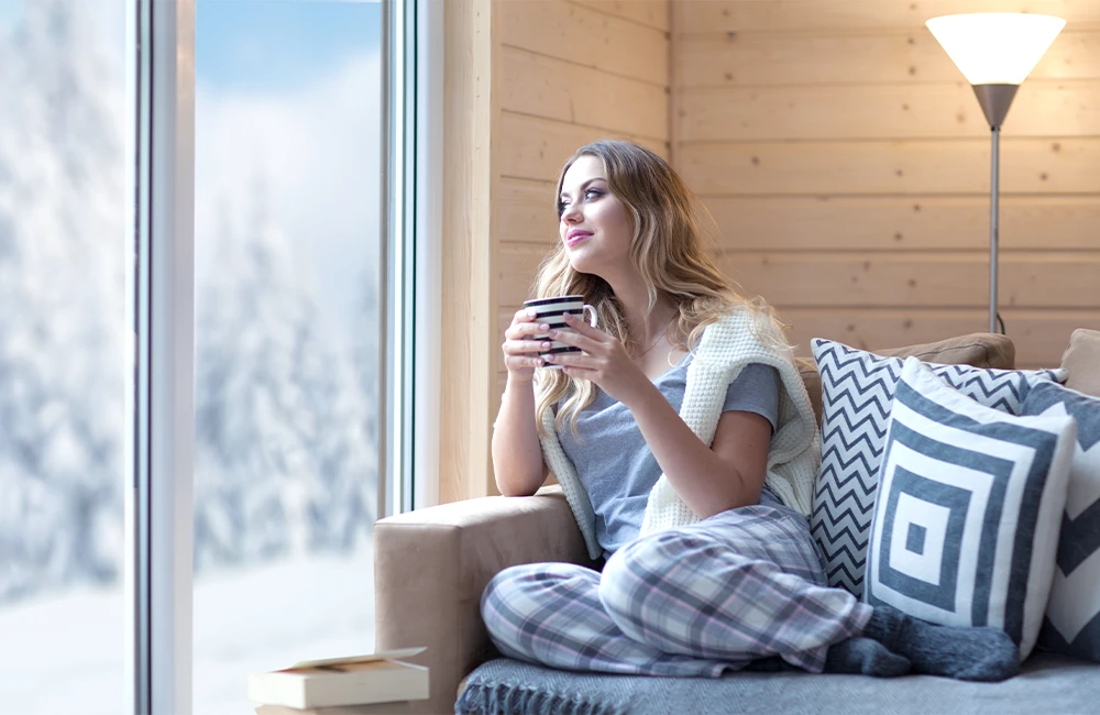 Une jeune femme installée dans son canapé, au chaud , regarde son jardin enneigé